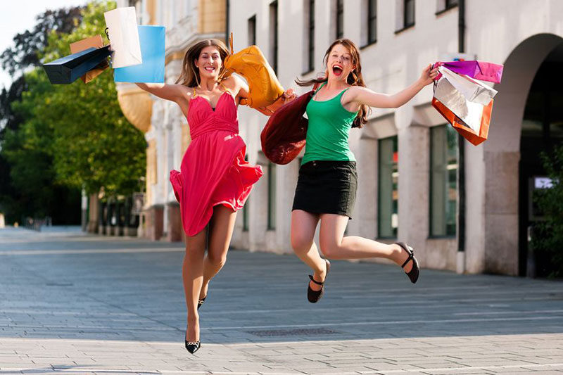 Zwei lächelnde Frauen springen freudig in die Luft und halten dabei mehrere bunte Einkaufstüten. Eine trägt ein rosa Kleid, die andere ein grünes Oberteil mit einem schwarzen Rock. Sie stehen an einem sonnigen Tag auf einem gepflasterten Weg mit Geschäften und Bäumen im Hintergrund.